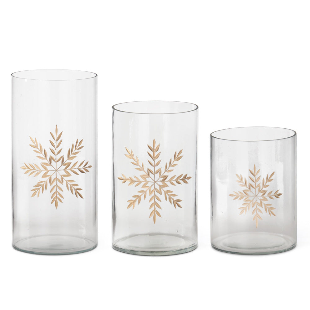 Three clear glass cylindrical containers featuring etched gold snowflake patterns in varying heights.
