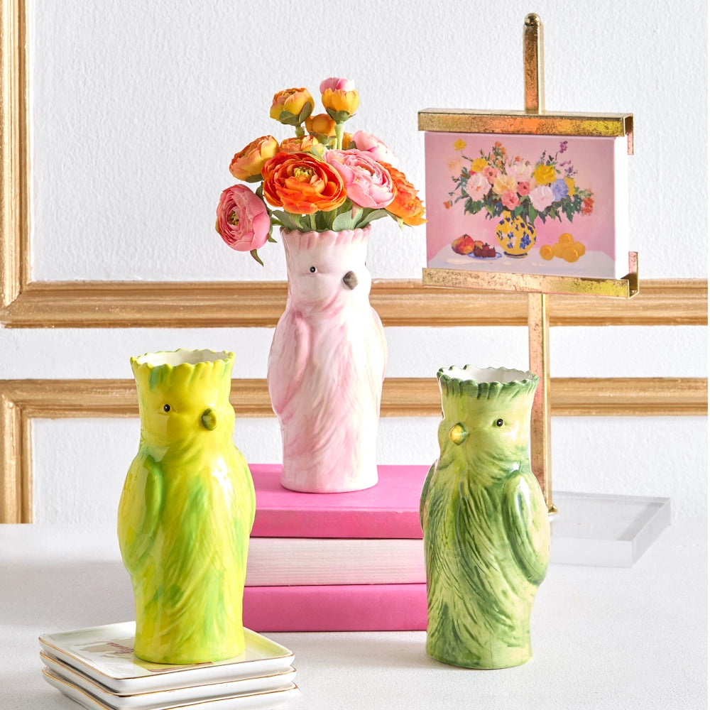 Ceramic vases shaped like birds, with green and pink hues, hold flowers. Bright blossoms in the background evoke a cheerful, artistic setting.