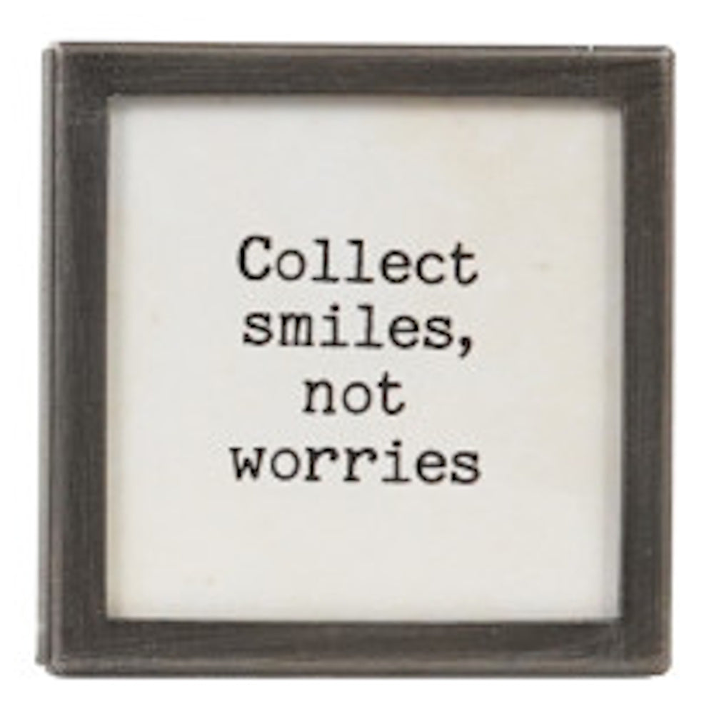 A square silver-framed sign with the text "Collect smiles, not worries" in black typewriter font.