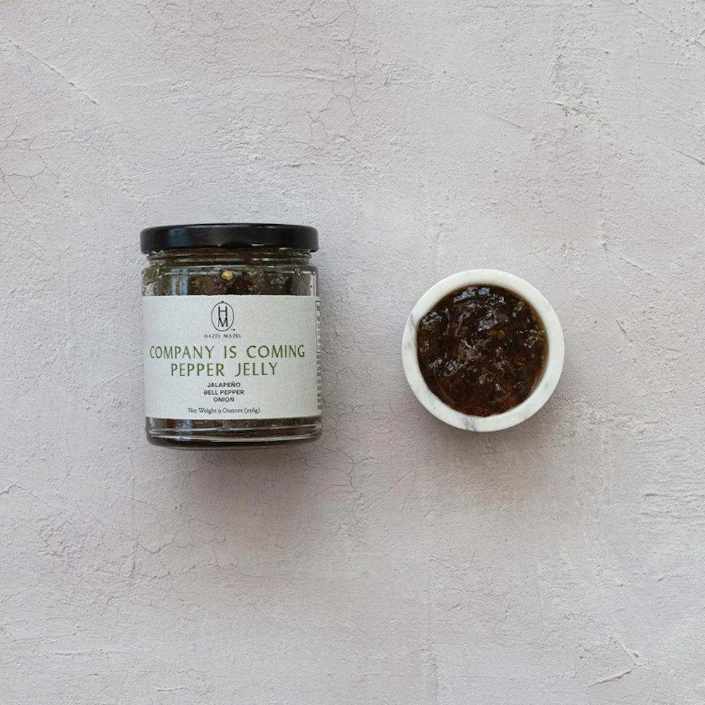 A jar of Hazel Hazel "Company Is Coming" Pepper Jelly next to a small white bowl of the dark, savory jam.