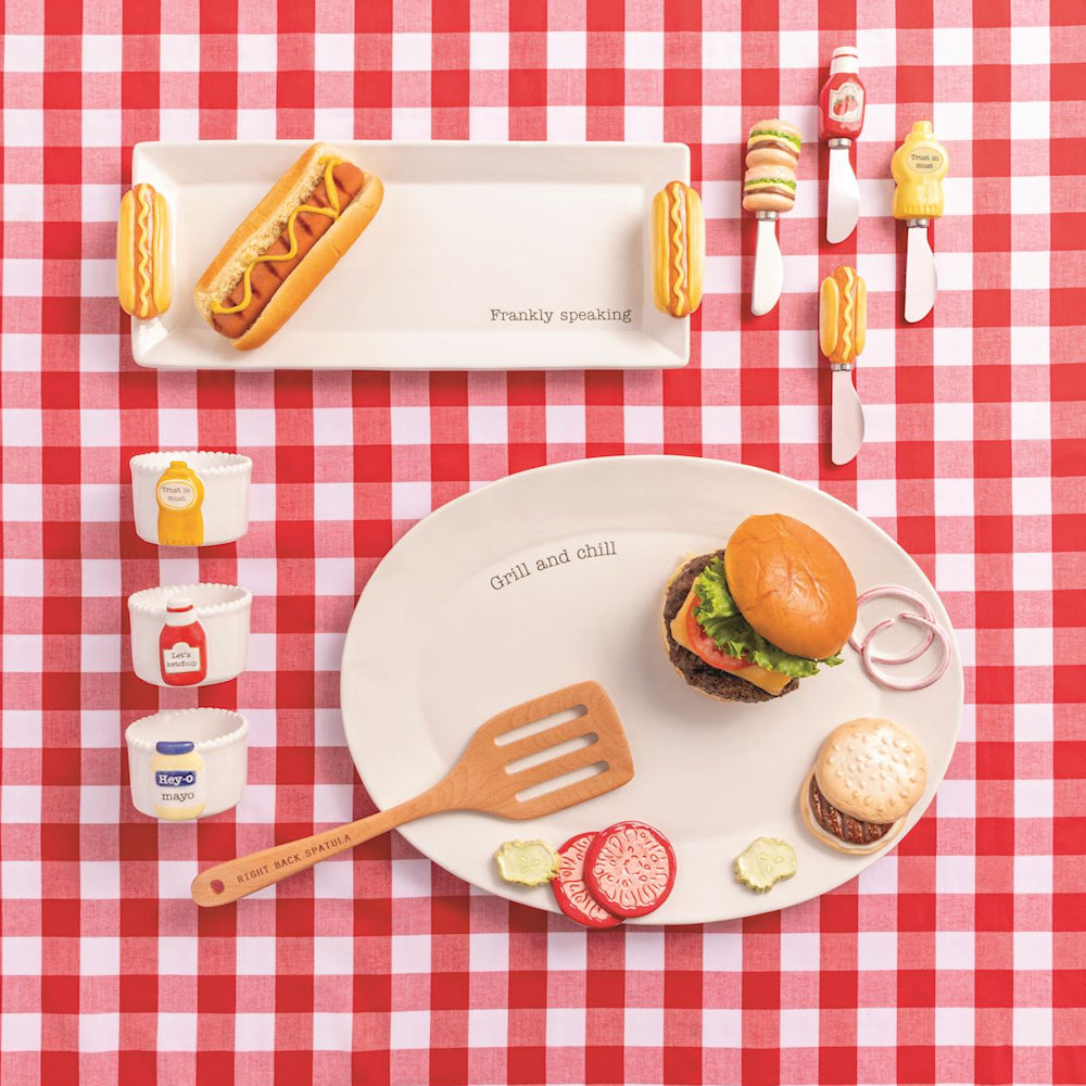 Gingham picnic table set with hot dog and burger platters, condiment-themed spreader knives, and ceramic dipping bowls.