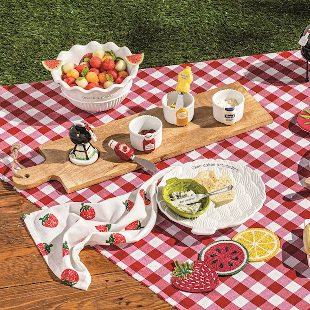 Whimsical picnic set with fruit-themed bowls, condiment servers, and a red checkered tablecloth on a sunny lawn.