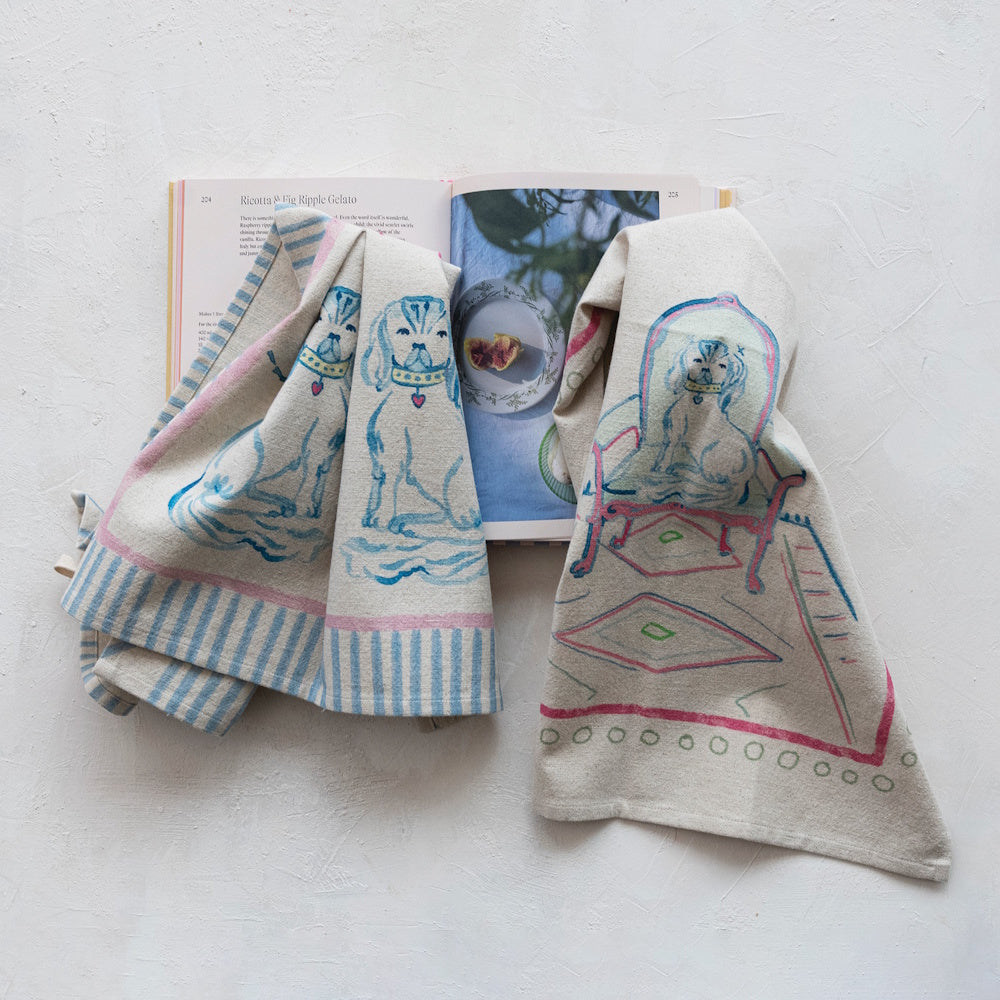 Two cotton tea towels with illustrated dog prints laid over an open cookbook on a white surface.