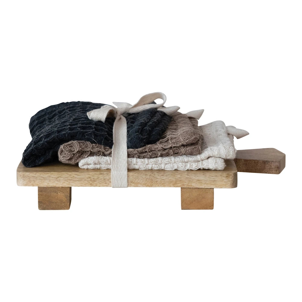 Stack of three textured cotton dish cloths tied with a ribbon on a footed mango wood serving board, isolated on white.