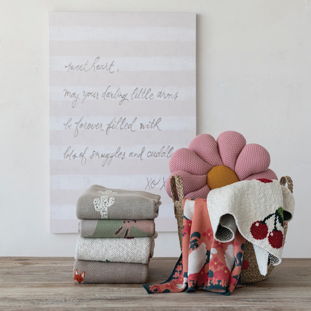 Folded and draped cotton knit baby blankets with swan, cherry, and cactus patterns next to a flower-shaped pillow.