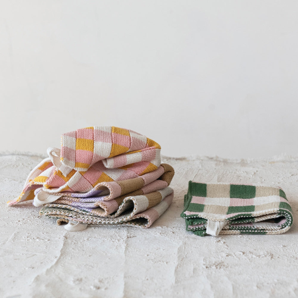 Stacked and folded checkered cotton knit dishcloths in multi-color patterns on a textured white surface.