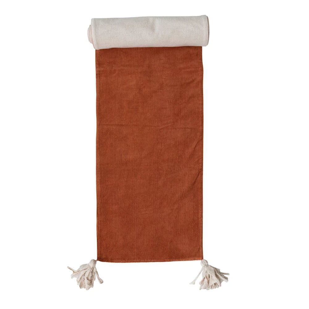 Terracotta corduroy table runner with white tassels and a rolled top edge on a white background.