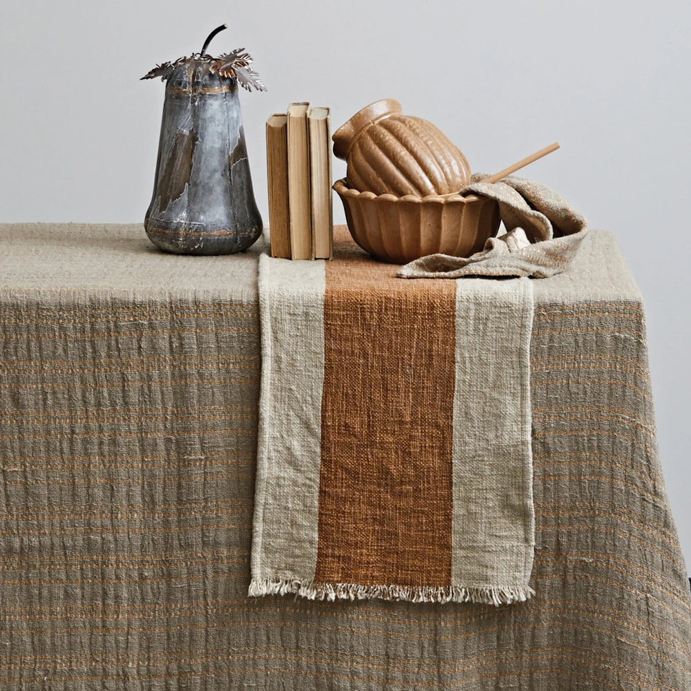 Fringed linen table runner with a thick terracotta center stripe draped over a rustic tablecloth.