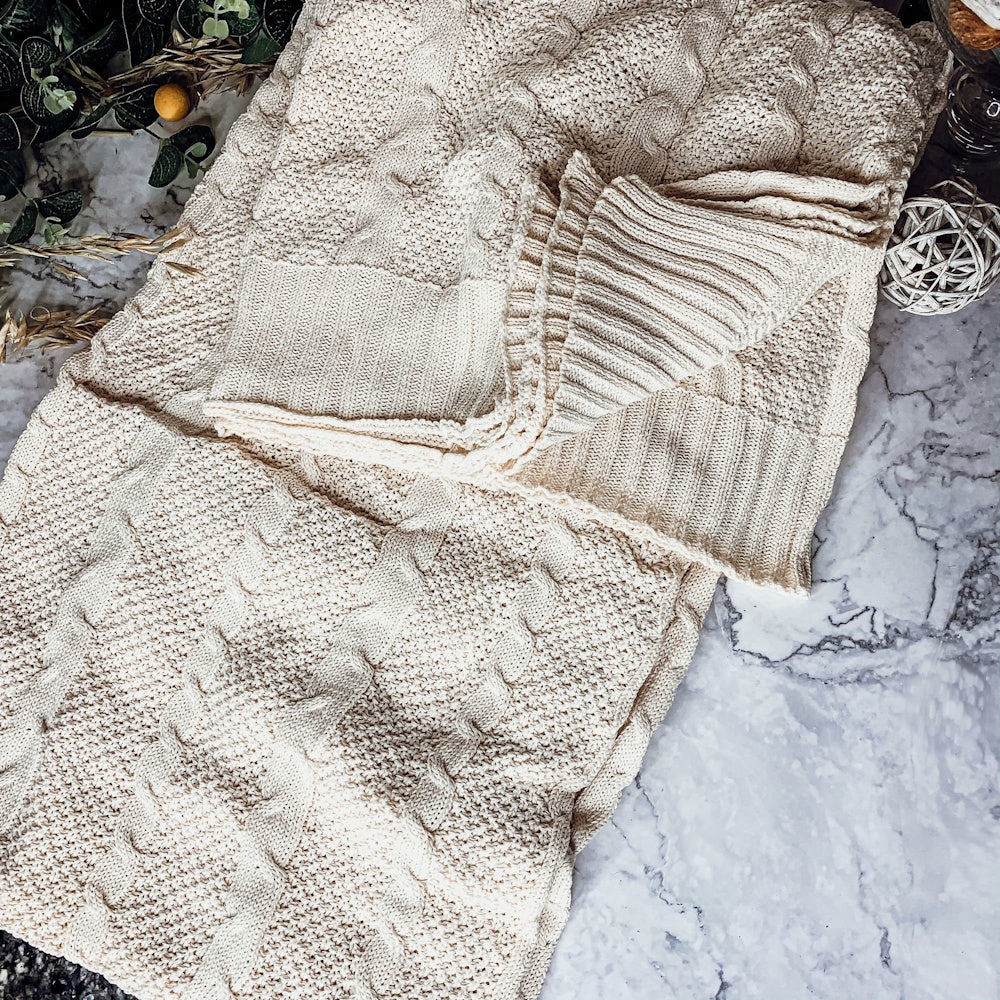 A folded cream-colored cable knit blanket resting on a marble surface.