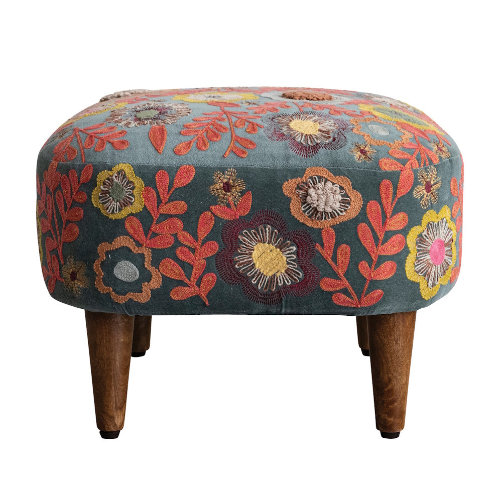 A blue velvet upholstered stool with colorful floral embroidery and short wooden legs against a white background.