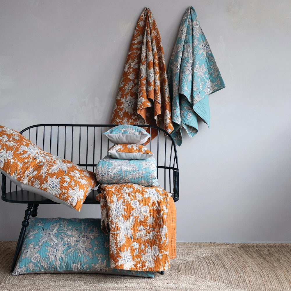 Hand-embroidered kantha stitch cotton voile throws and pillows with floral patterns on a black bench and wall.