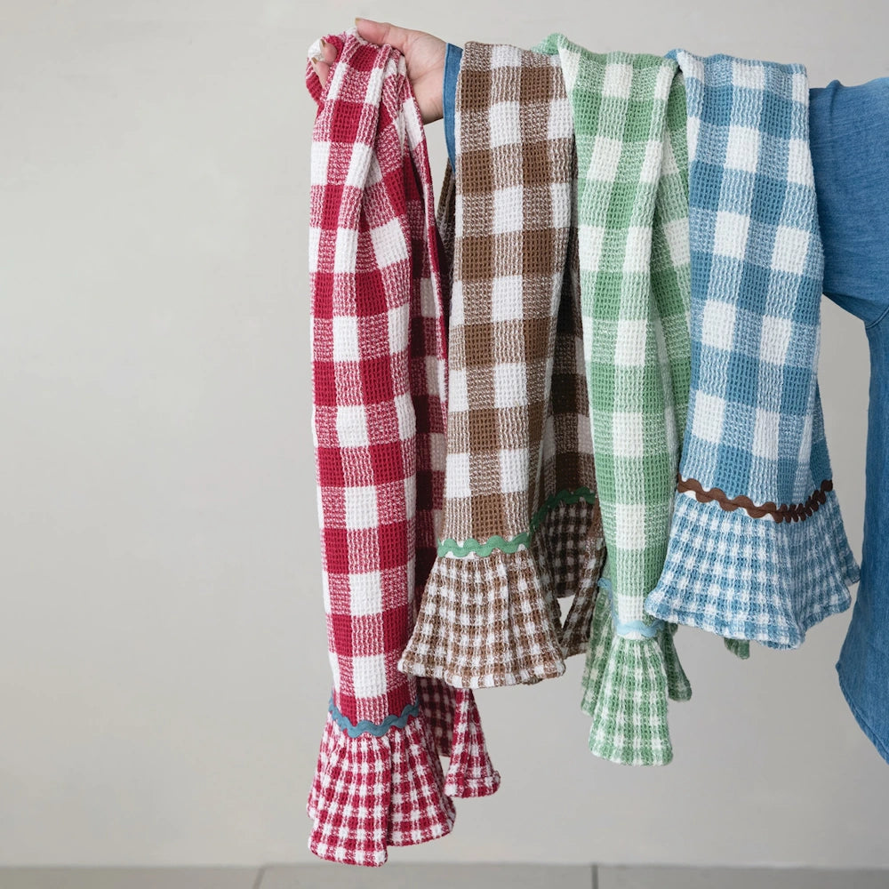 Hand holding four buffalo check waffle-weave tea towels in red, brown, green, and blue with ruffled hems.