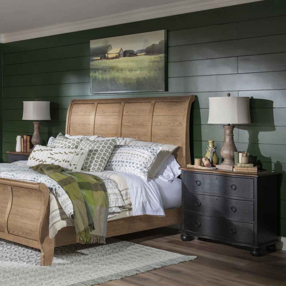 Bedroom with black three drawer accent chest used as a nightstand beside a wood sleigh bed and lamp.