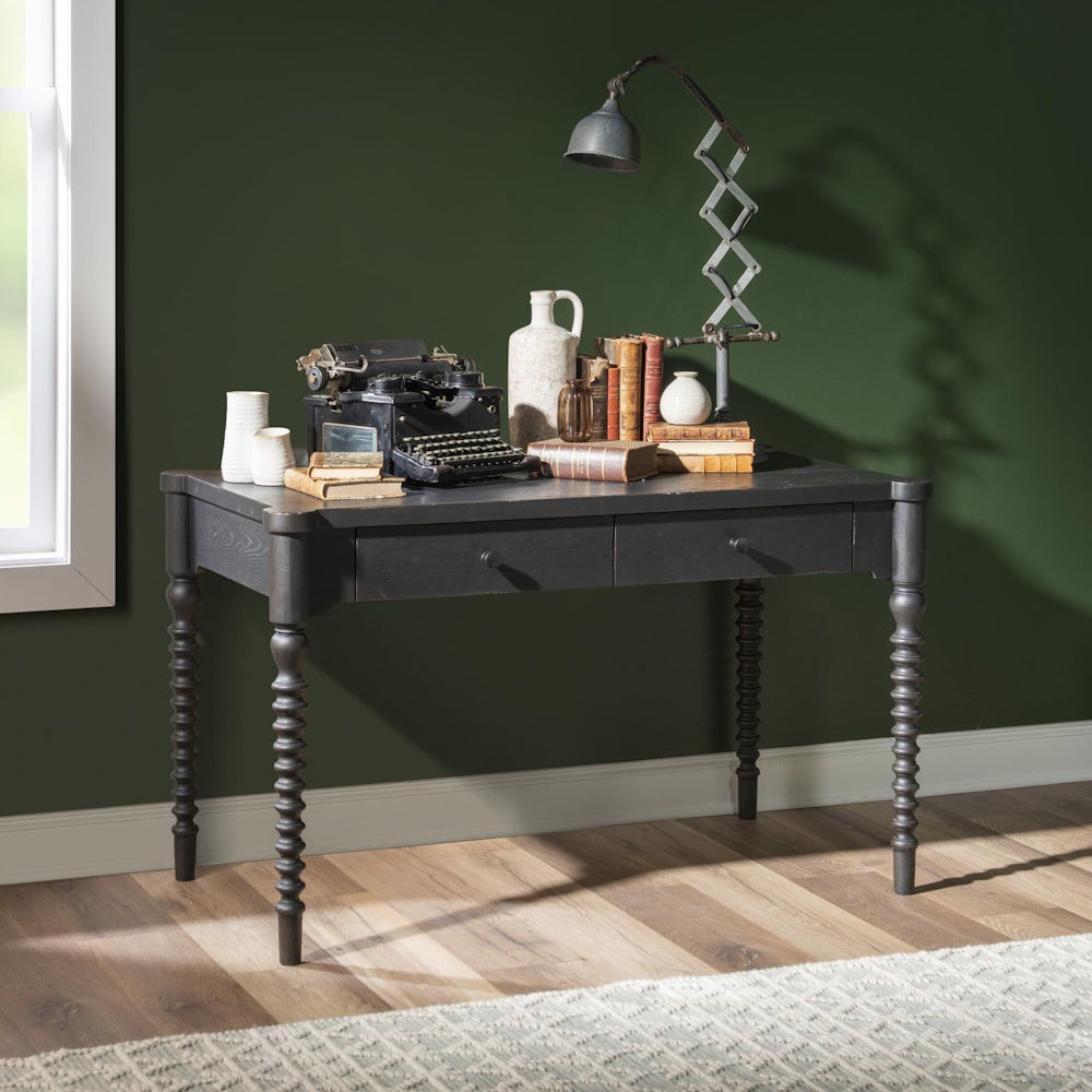 Dark bobbin-leg desk styled with a vintage typewriter, books, and lamp against a deep green wall.