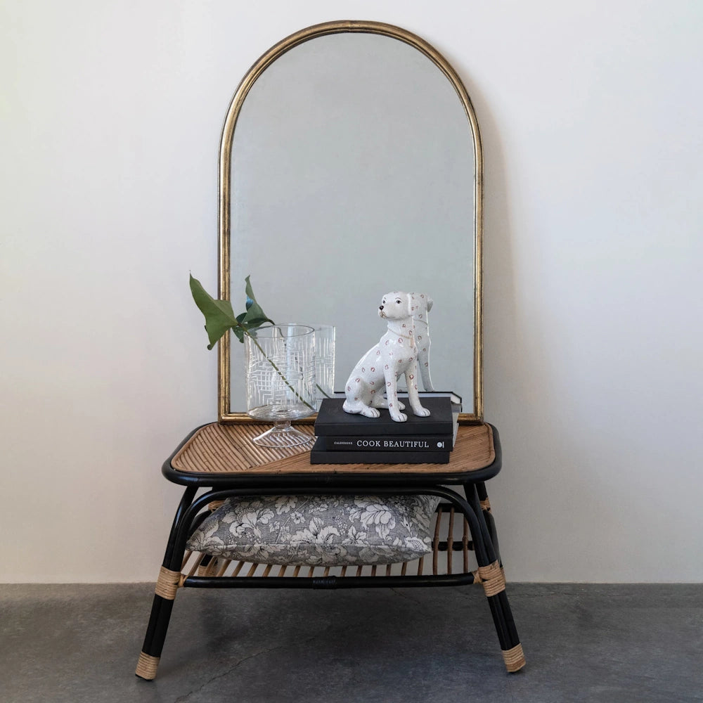 Hand-painted ceramic dog figurine on black books in front of an arched gold mirror on a rattan side table.