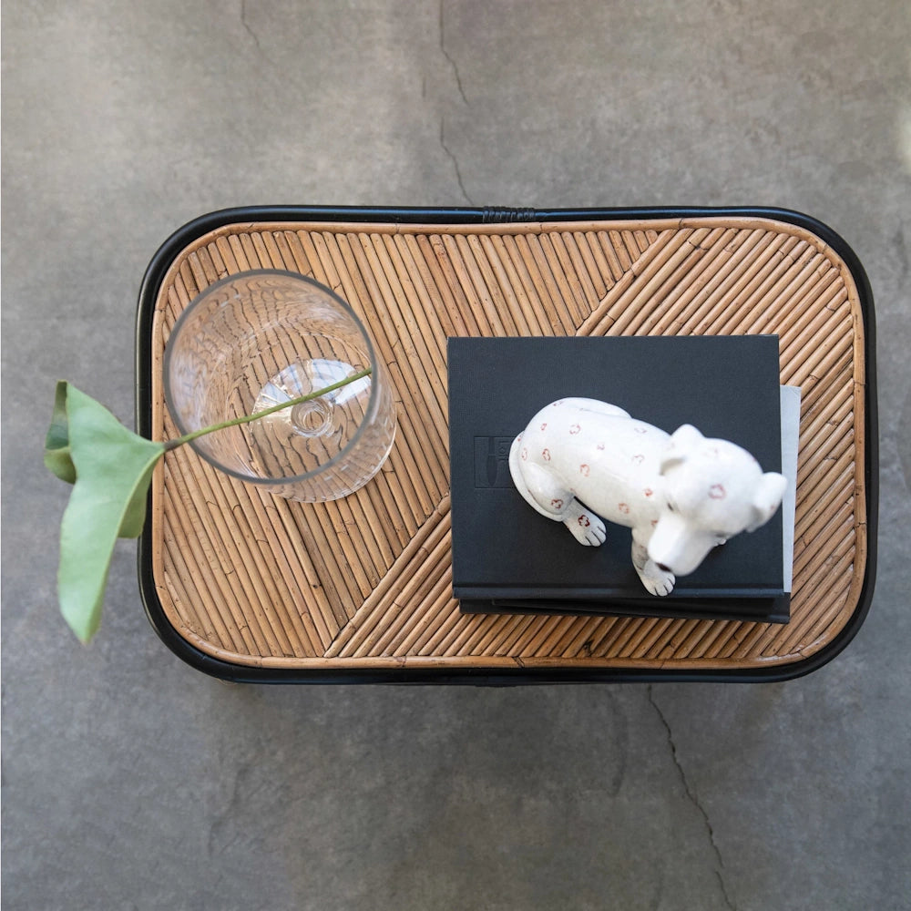 Top view of a rattan coffee table with a glass vase, green leaf, and a white ceramic dog figurine on black books.