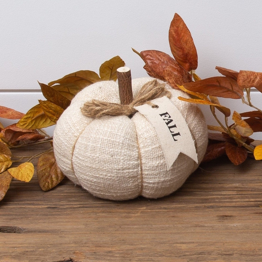 Cream fabric pumpkin with a wooden stem, twine bow, "FALL" tag, and autumn leaves on wood.