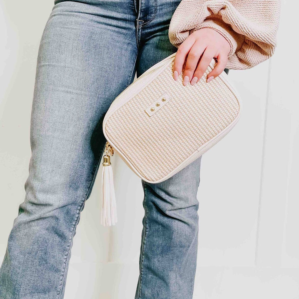 A person wearing blue jeans holds a beige textured clutch purse with a white tassel zipper pull.