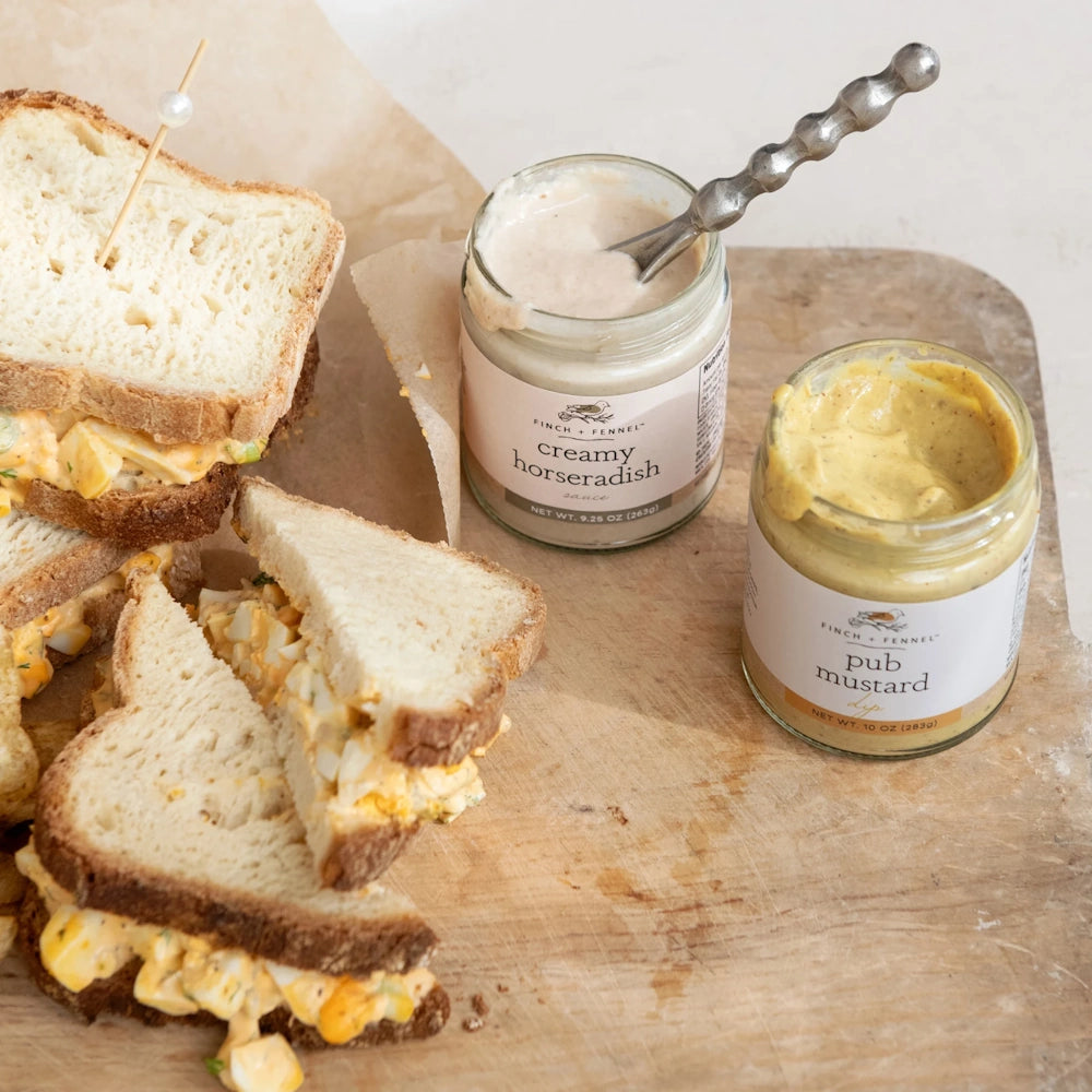Egg salad sandwich halves on a wooden board next to open jars of Finch + Fennel horseradish sauce and pub mustard.