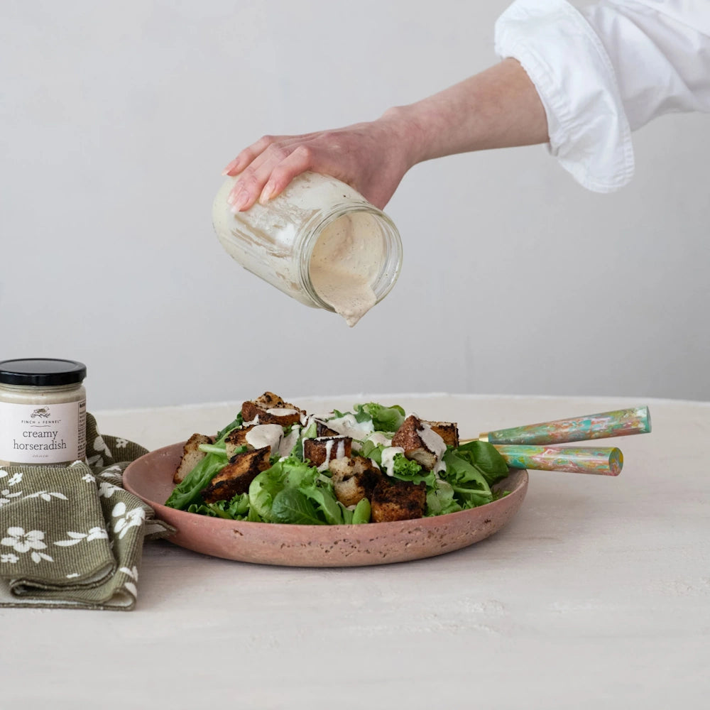 A hand pouring creamy dressing from a glass jar onto a fresh green salad with croutons in a pink ceramic dish.