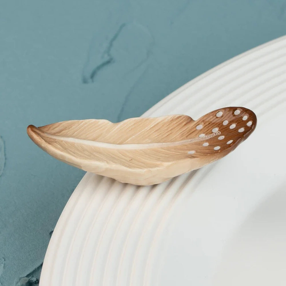 Ceramic feather-shaped dish with brown and white polka-dot patterns resting on the edge of a white plate.