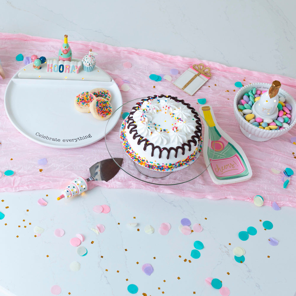 Festive birthday party table with white sprinkle cake, "Hooray" serving plate, cookies, and colorful confetti on pink runner.