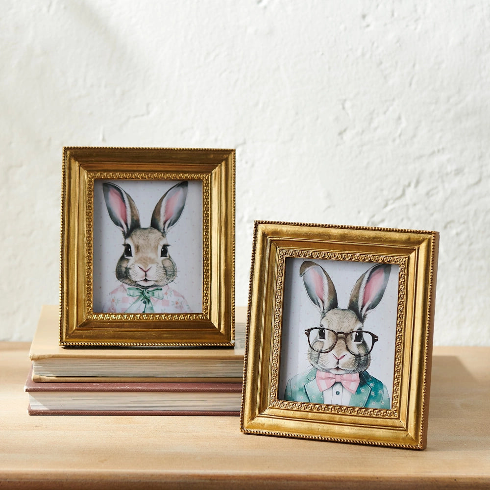 Two whimsical framed illustrations of rabbits in elegant attire, displayed on a stack of books against a textured wall.