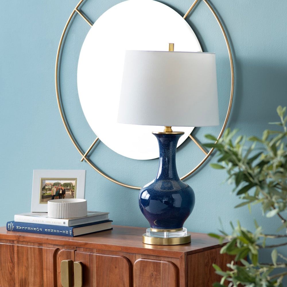 Modern blue ceramic table lamp with white shade on a wooden console table against a blue wall with a round gold mirror.