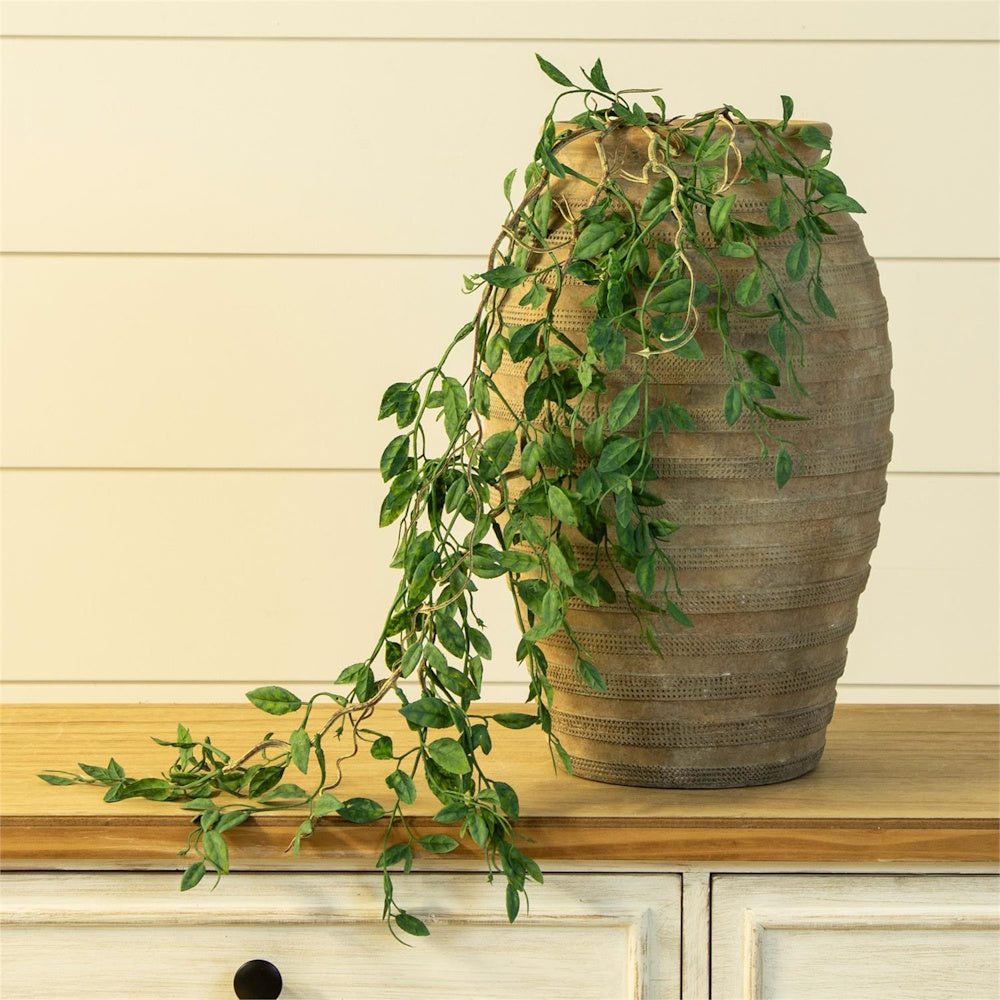 Draping faux green leaves in a rustic textured terracotta vase on a wooden sideboard with a cream shiplap background.