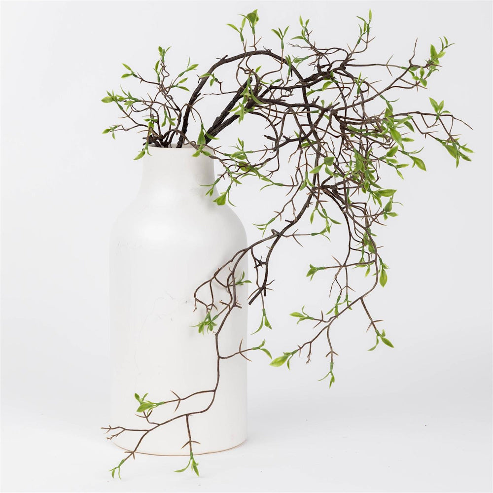 Artificial early spring vine stem with green leaves cascading from a modern white ceramic vase on a white background.