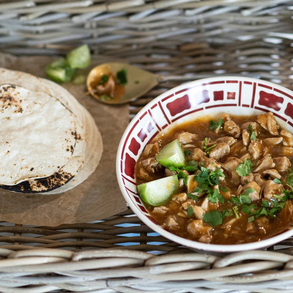 Bowl of pork carnitas with lime and cilantro, served with a side of toasted corn tortillas on a woven surface.