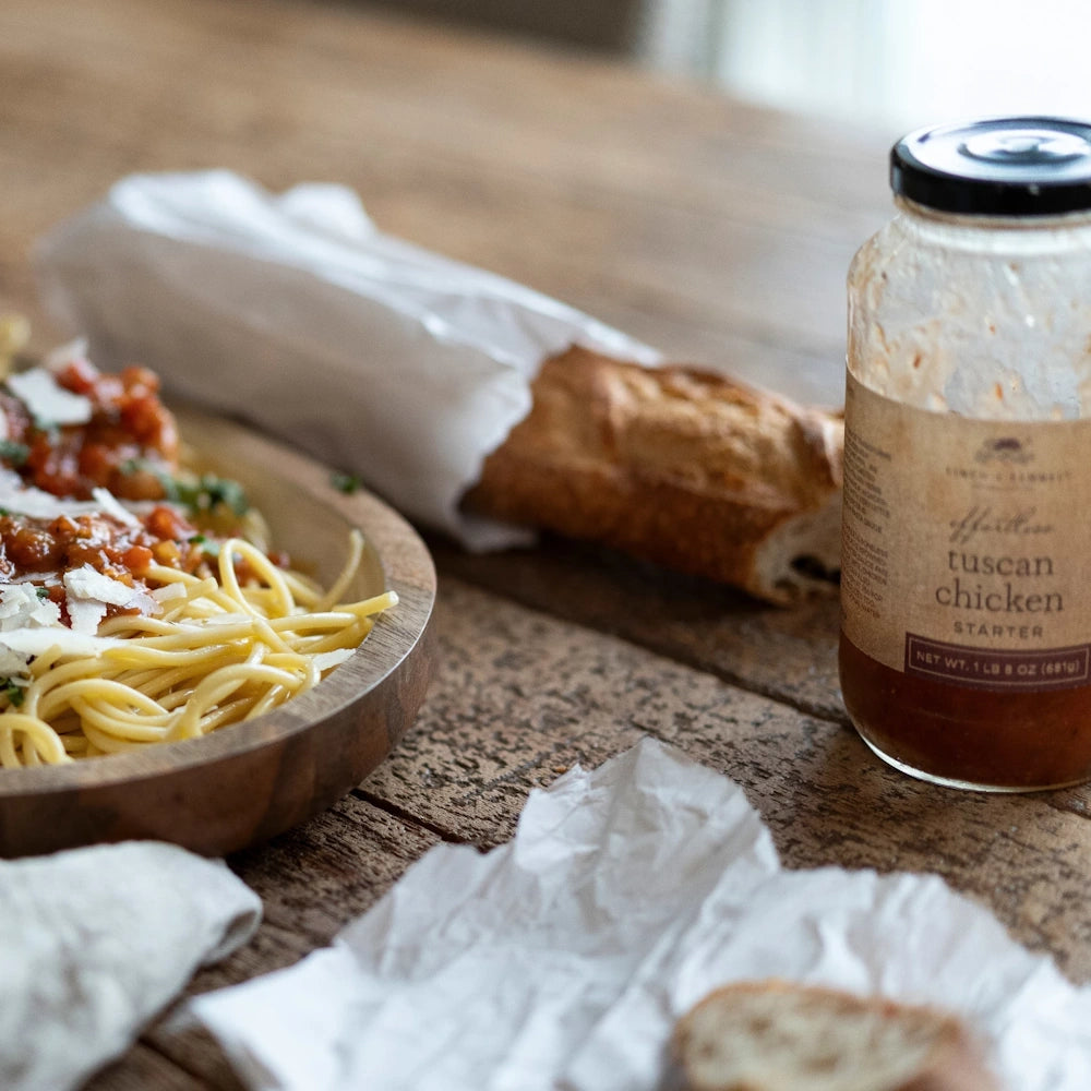 Tuscan chicken pasta in a wooden bowl next to a crusty baguette and a jar of Tuscan chicken starter sauce on a wood table.