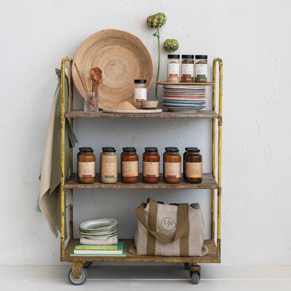 A rustic three-shelf rolling cart displays assorted sauce jars, spice containers, plates, and a branded canvas tote bag.