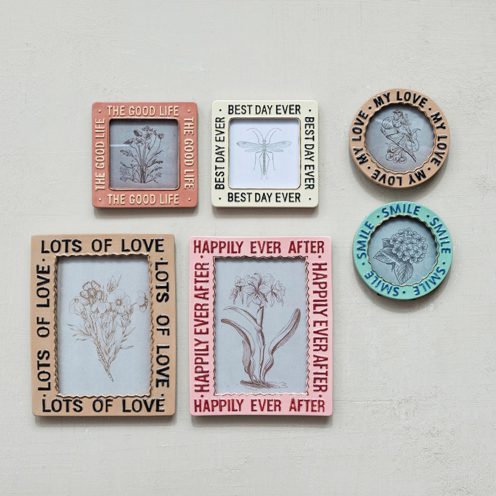 Six colorful, embossed stoneware photo frames with phrases and botanical sketches hanging on a plain wall.