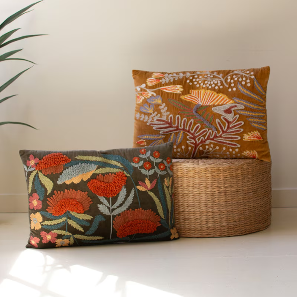 Two flower-embroidered pillows in brown and dark grey styled next to a woven ottoman.