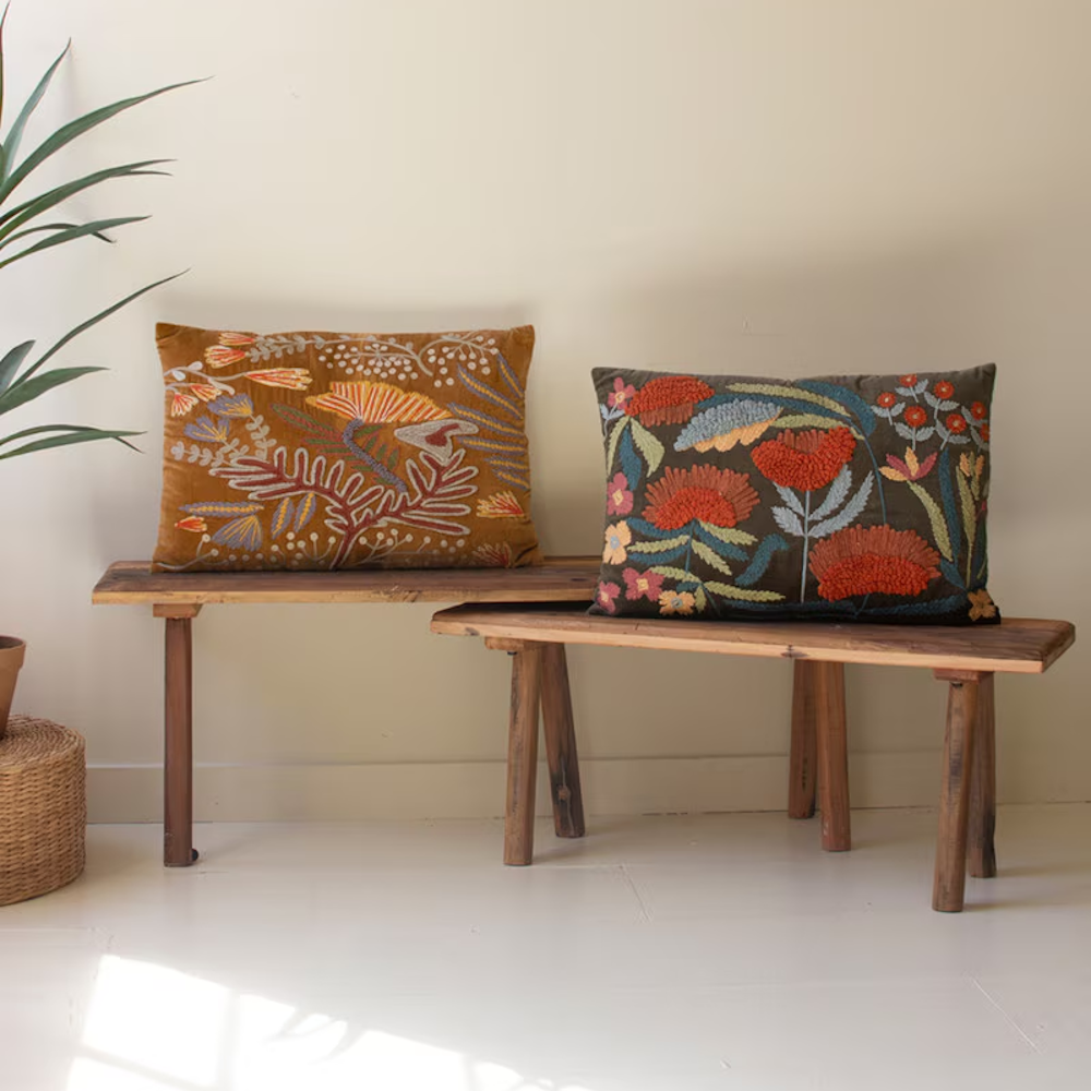 Two embroidered floral throw pillows displayed on rustic wooden benches against a cream wall.