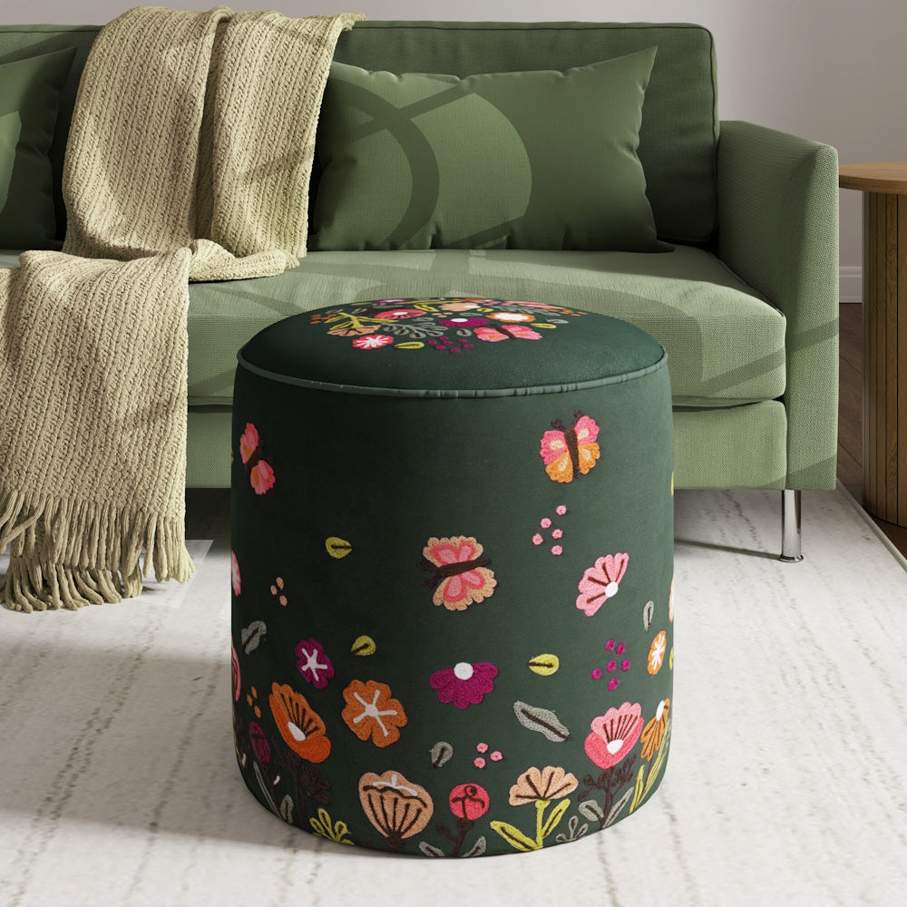 Dark green cylindrical ottoman with embroidered colorful flowers and butterflies in front of a green sofa.