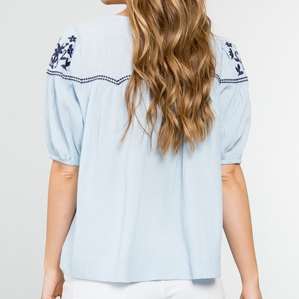 Back view of a light blue short-sleeve top with navy blue floral embroidery on the shoulders.