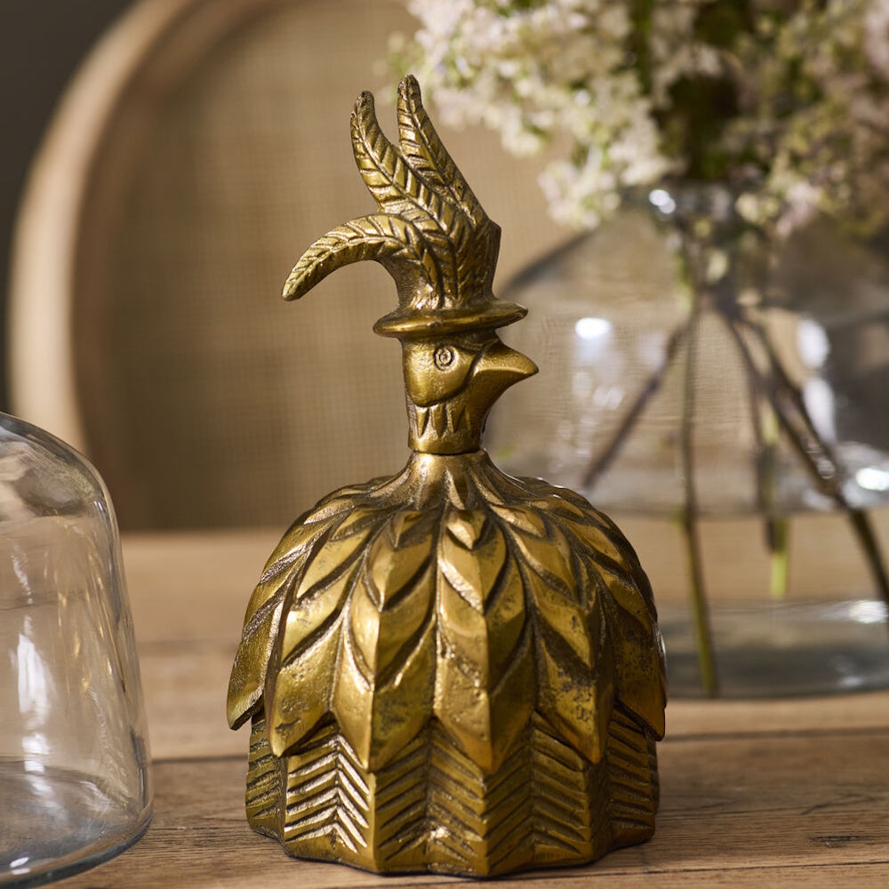 Antique gold decorative box shaped like a bell with a feathered bird top hat handle, displayed on a wooden table near a vase of flowers.