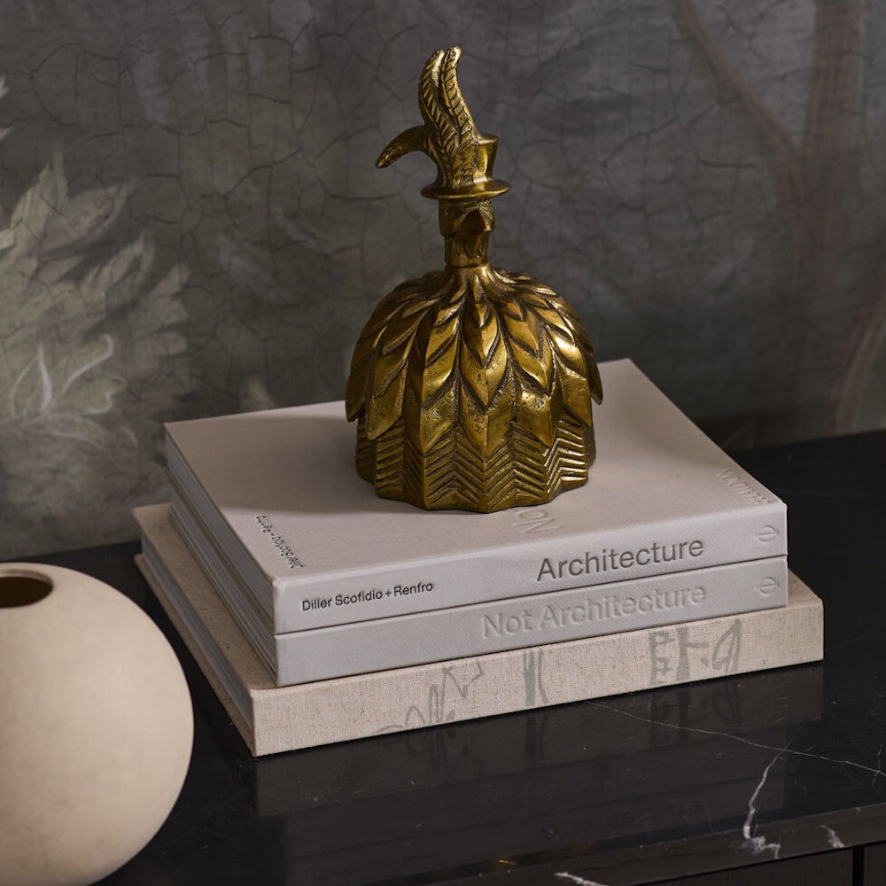 Antique gold decorative box shaped like a bell with a feathered bird top hat handle, displayed on top of a stack of architecture books.
