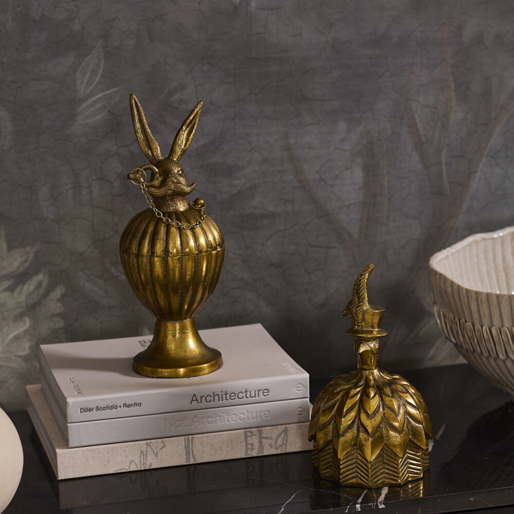 Antique gold decorative box shaped like a pedestal urn with a mustached rabbit lid next to a bell-shaped decorative box with a feathered bird top hat handle on a tabletop.