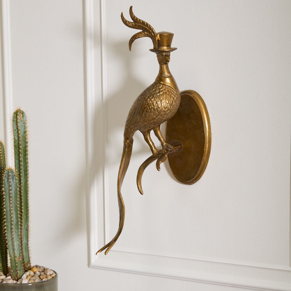 Brass bird wall sculpture wearing a top hat with a long feather crest and tail, mounted on a white wall.