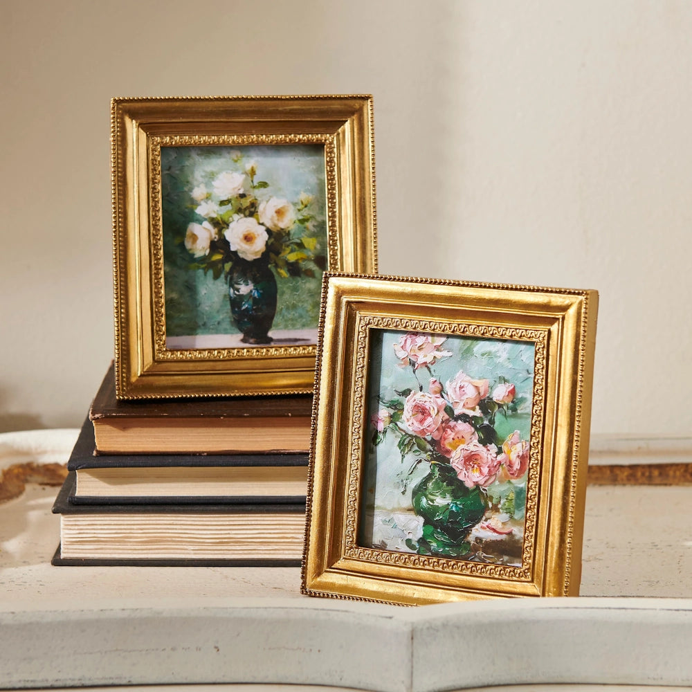 Two gold-framed floral paintings, one with white flowers in a dark vase and another with pink flowers, sit atop stacked books.