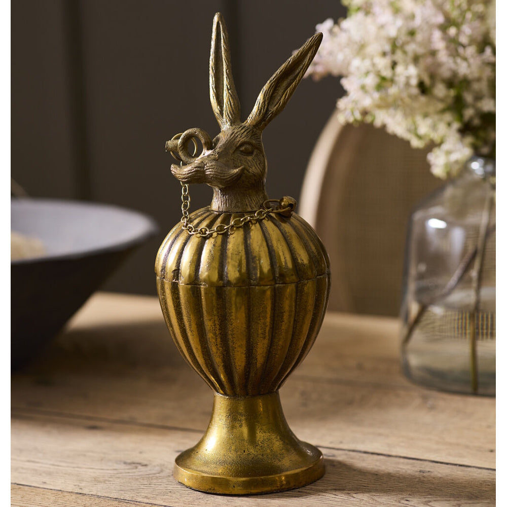 Antique gold decorative pedestal urn box with a mustached rabbit lid, displayed on a wooden table with a vase of flowers.