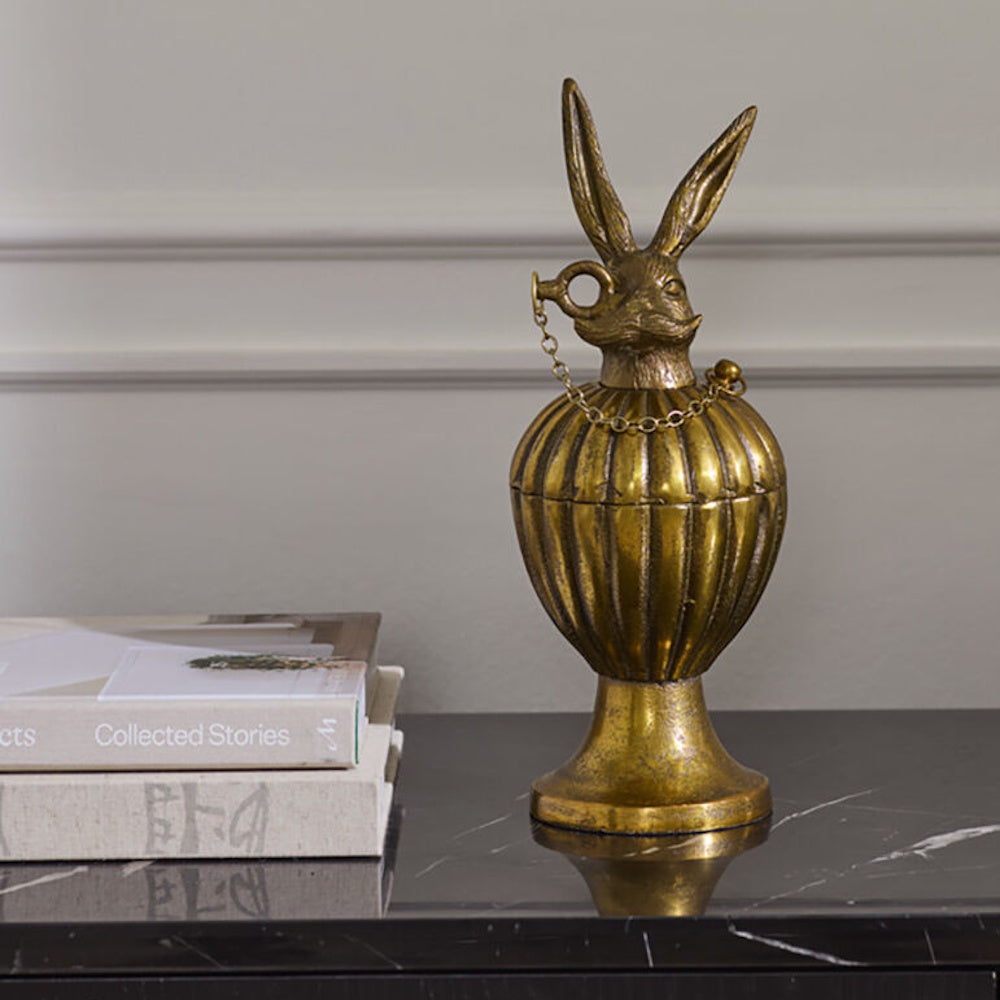 Antique gold decorative pedestal urn box with a mustached rabbit lid, displayed next to a stack of books on a dark tabletop.