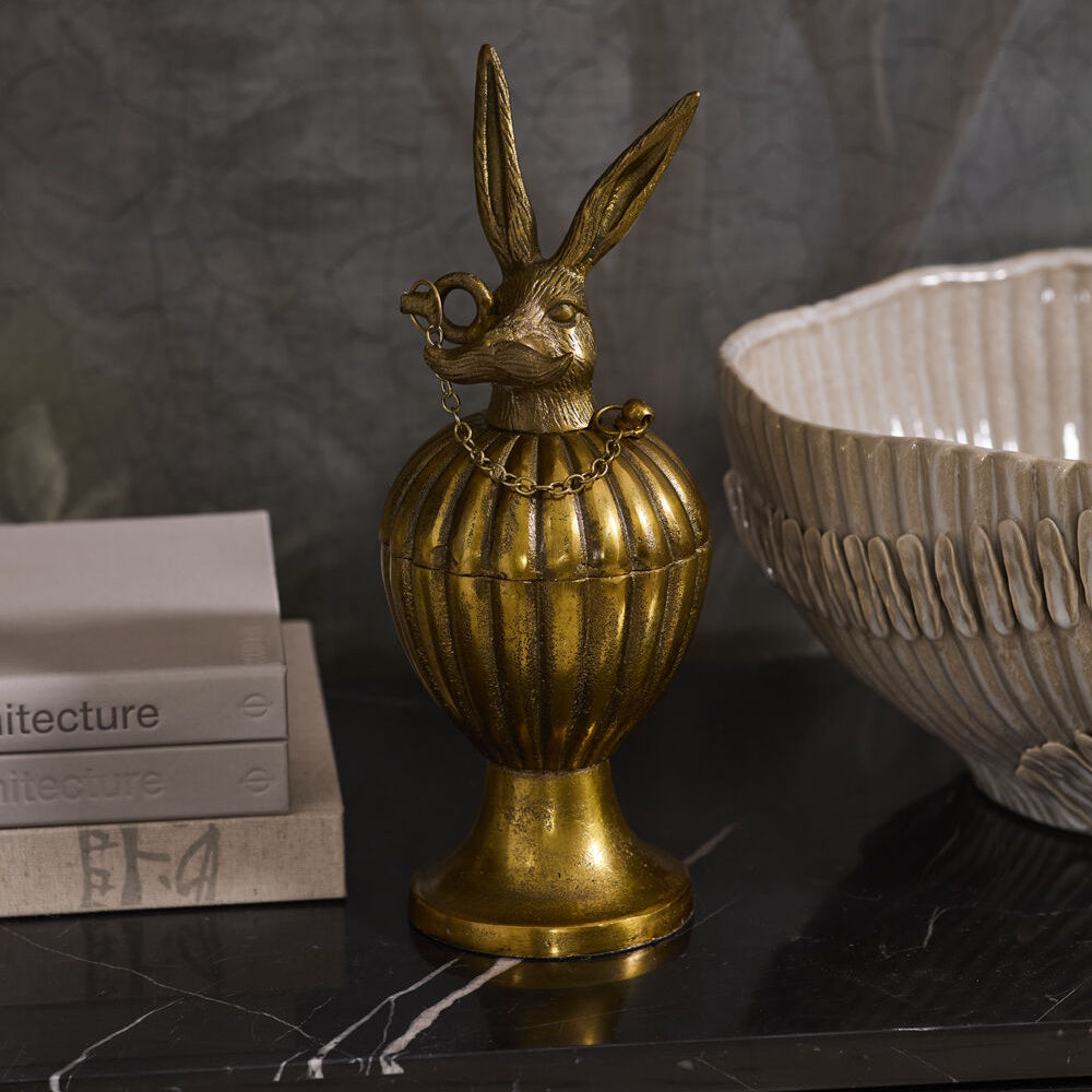 Antique gold decorative pedestal urn box with a mustached rabbit lid, displayed next to books and a large bowl on a dark surface.
