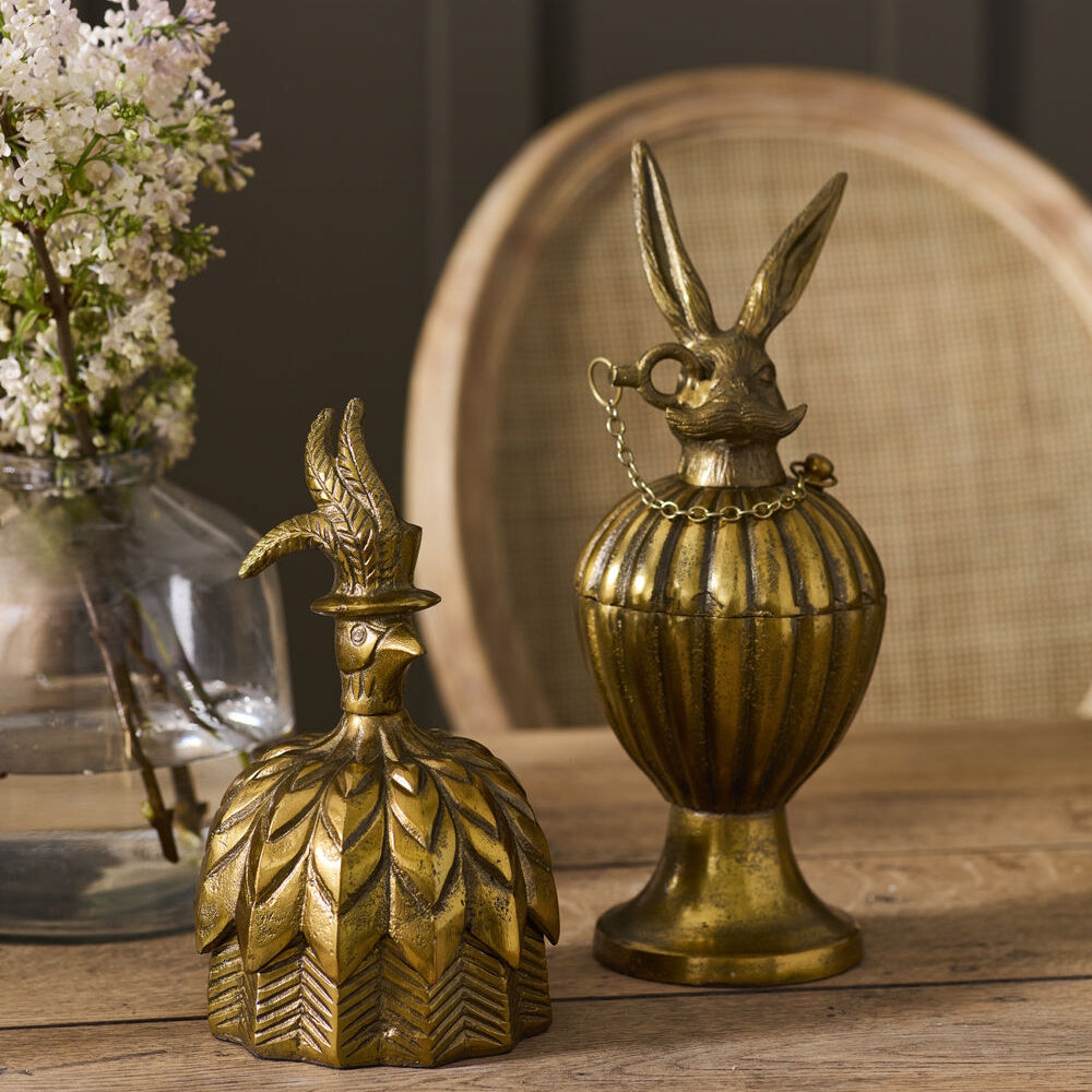 Antique gold decorative pedestal urn box with a mustached rabbit lid displayed alongside an ornate feathered bird figure on a wooden table.