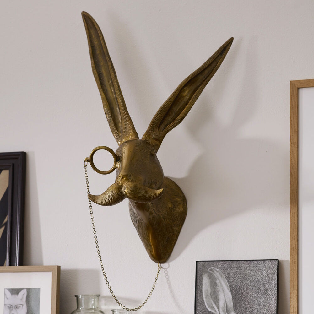 Brass wall mount of a hare's head with long ears, a mustache, and a monocle on a chain.