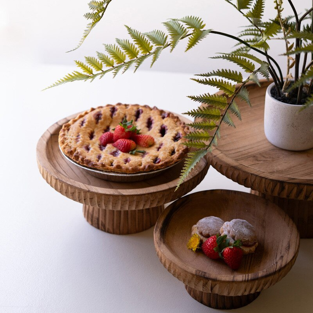 Three carved wooden pedestal stands holding a berry pie, pastries, and a potted fern on white.