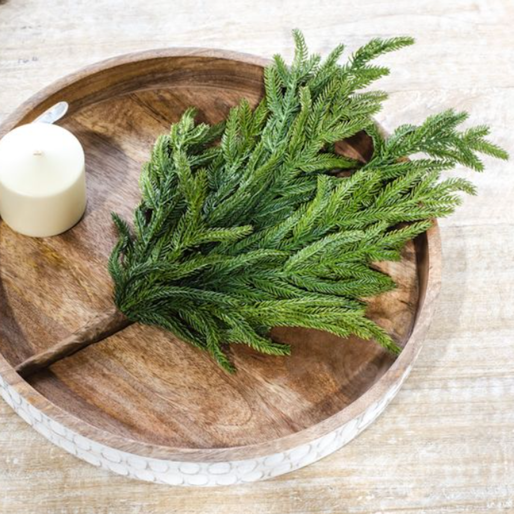 A realistic faux evergreen pine branch resting on a round wooden tray next to a white pillar candle.